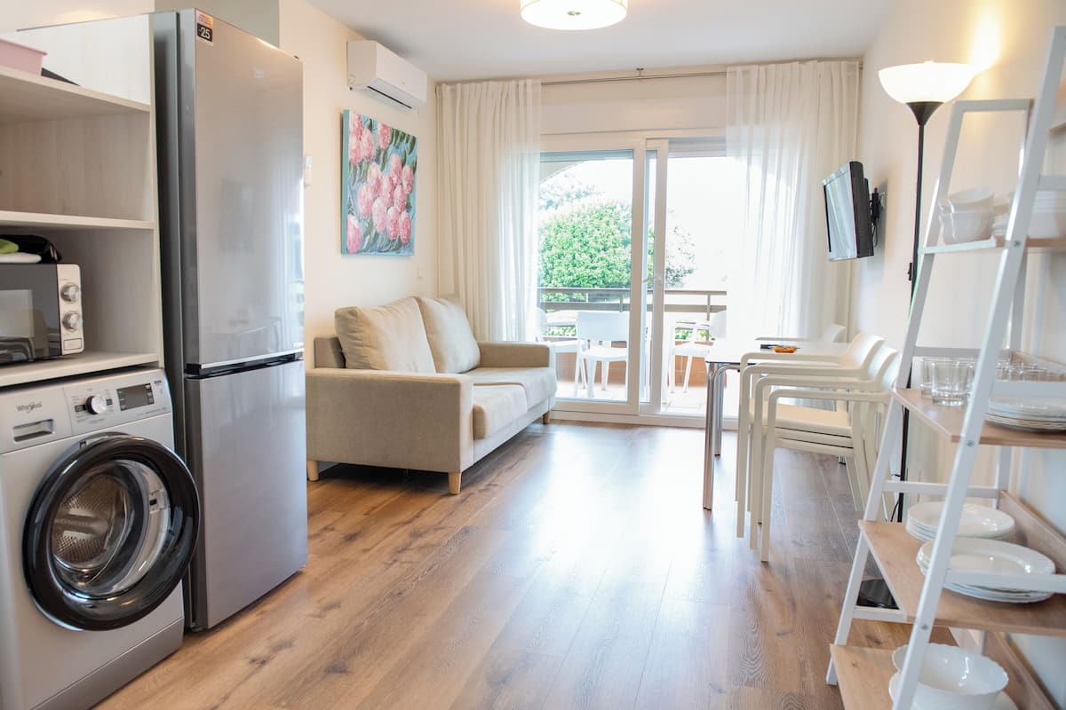 A compact living space with a washing machine, a sofa, a dining table with chairs, and a TV mounted on the wall.
