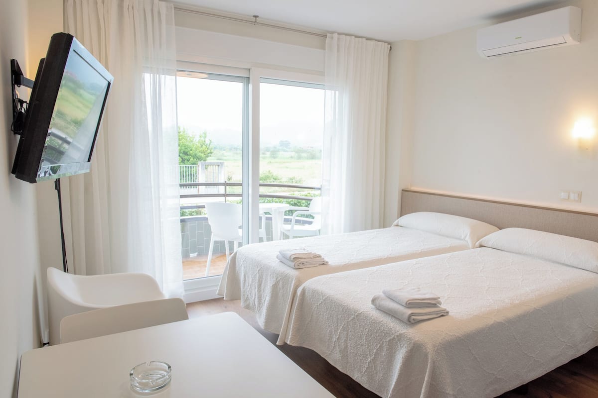 A hotel room with two beds, a TV, a table, and a balcony with a view of greenery.