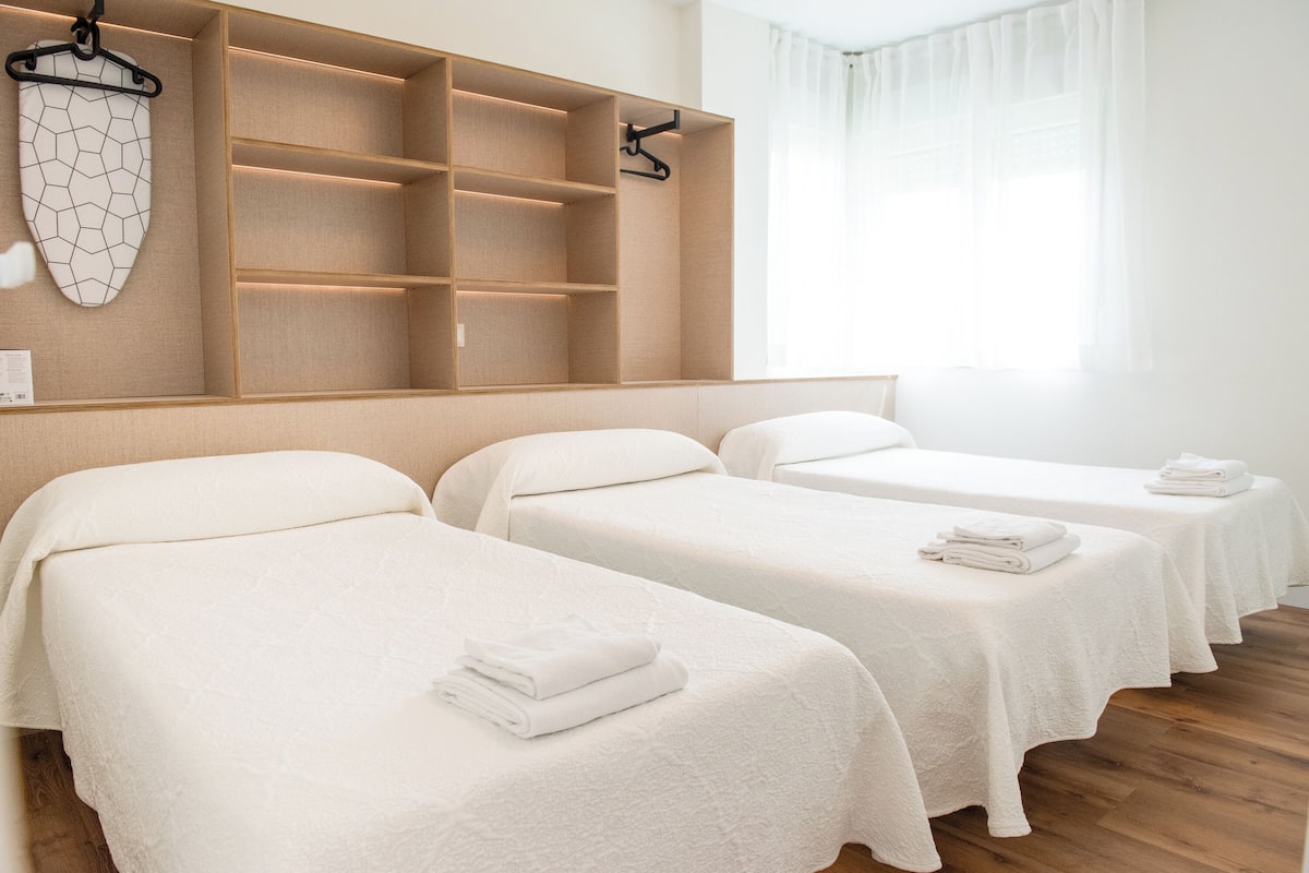A hotel room with two beds, a wooden shelf unit, and a window with sheer curtains.