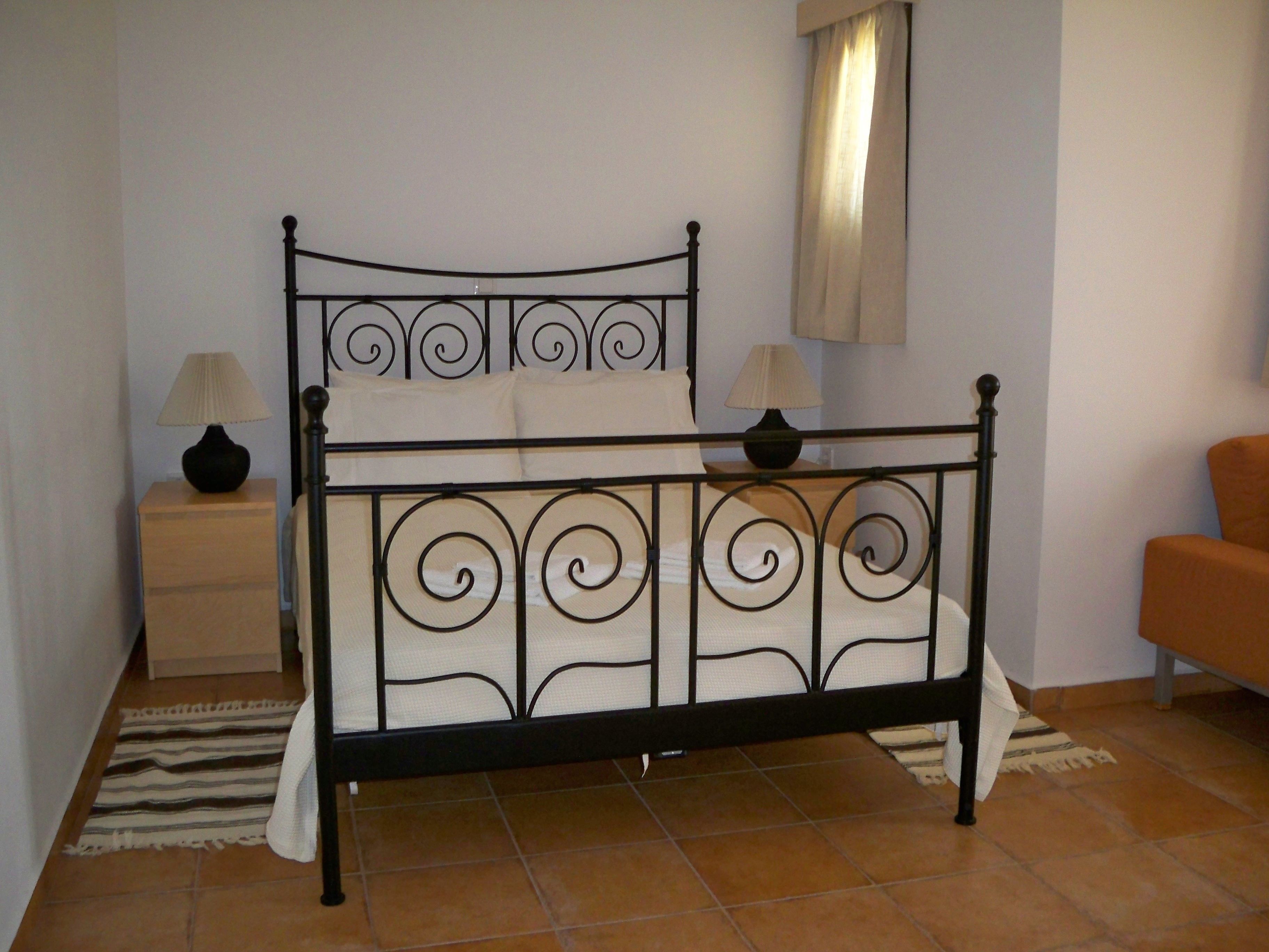 A bed with a metal frame and white bedding, a nightstand with a lamp, and a chair in a room with tiled flooring.