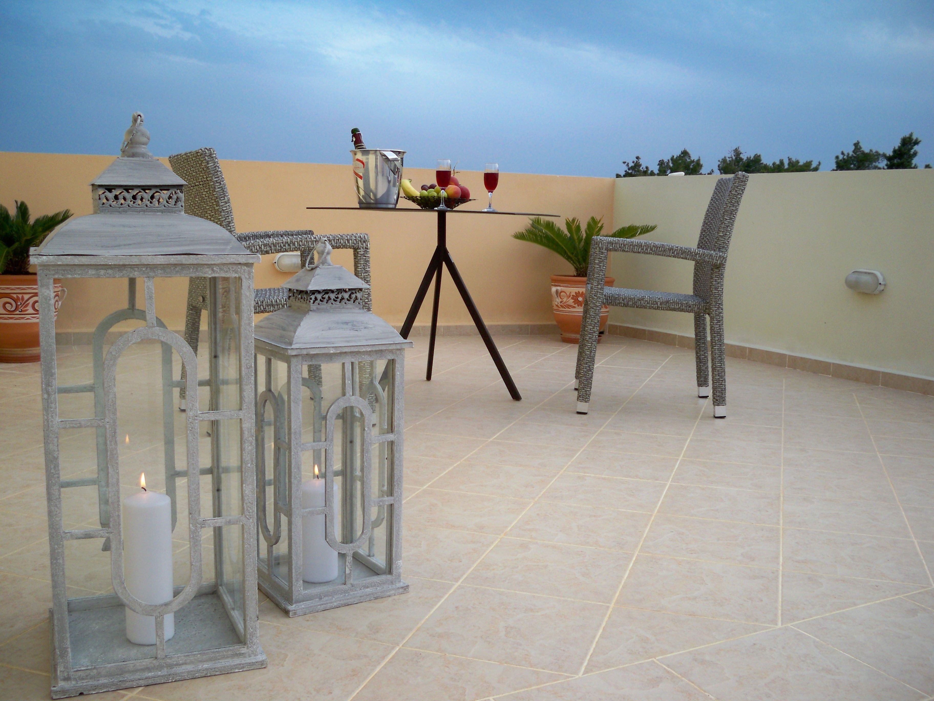 Eine Terrasse im Freien mit einem für zwei Personen gedeckten Tisch, mit Obstschale, einer Flasche und einem Glas Rotwein. Es sind Korbstühle und zwei Laternen mit Kerzen vorhanden.
