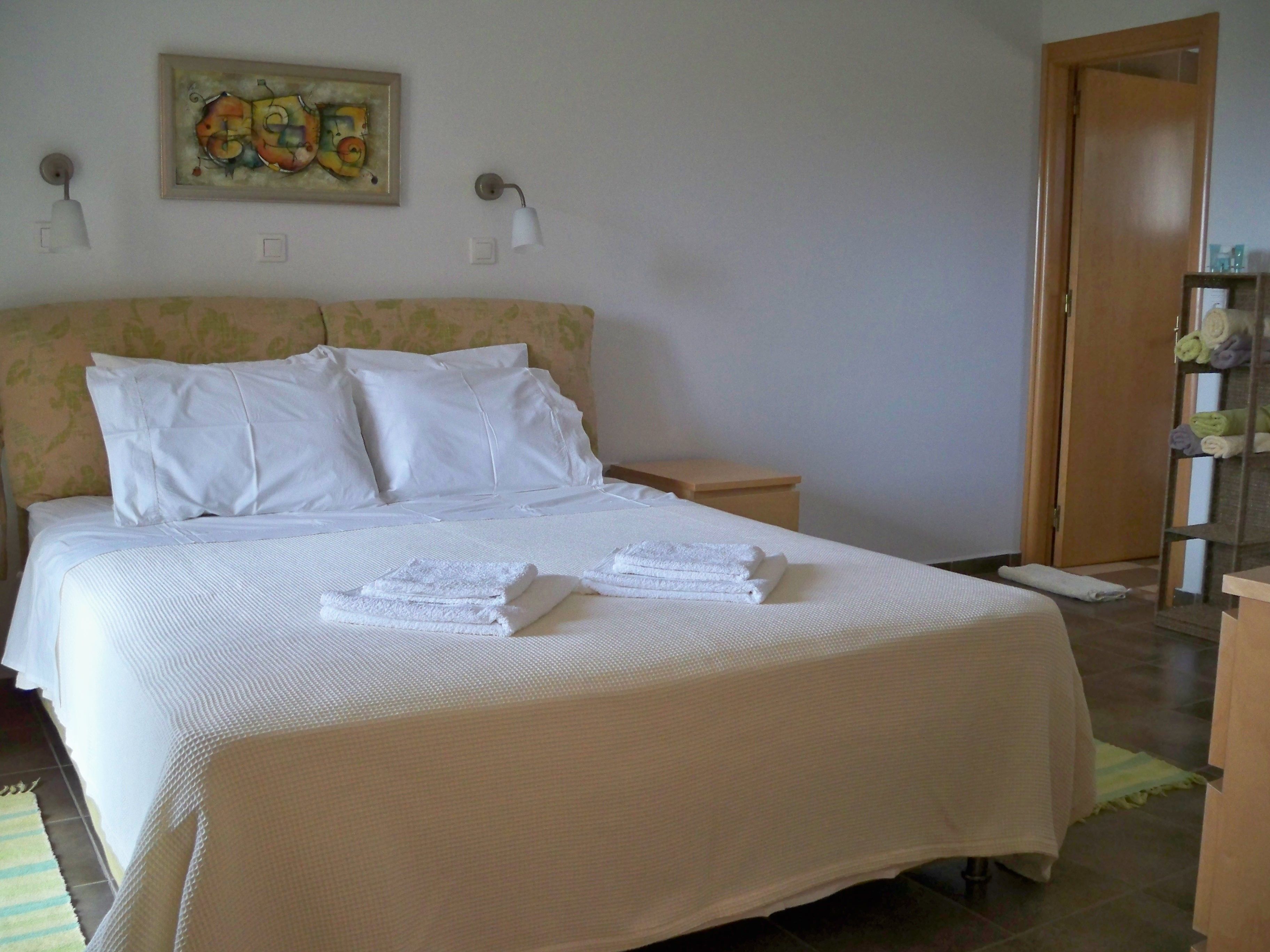 A neatly made bed with white linens and towels, a small bedside table, a wall-mounted lamp, and a door to the right.