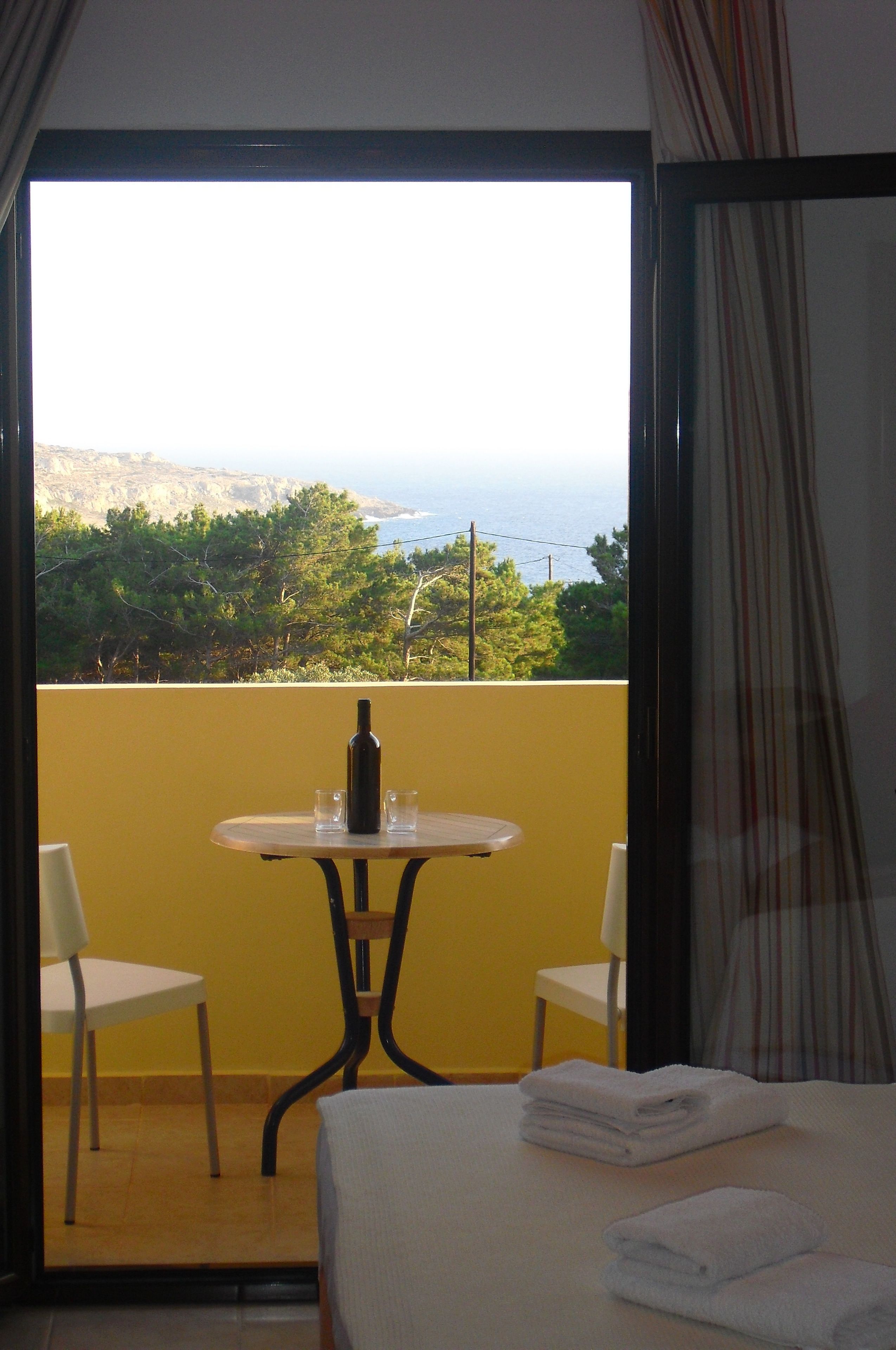 A room with a bed, a table with a bottle and glasses, two chairs, and a view of the sea and mountains.
