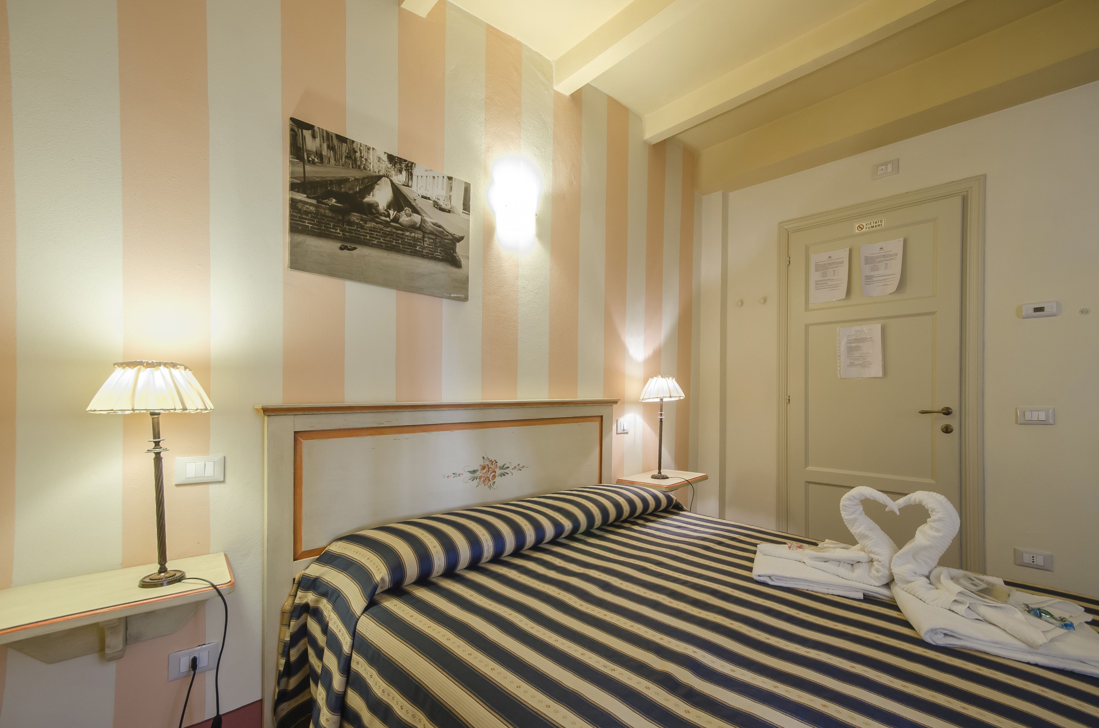 A hotel room with striped bedding, a headboard with floral decoration, two bedside lamps, and a door with papers on it.