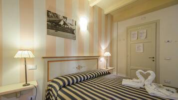 Habitación de hotel con ropa de cama a rayas, cabecera decorada con motivos florales, dos lámparas de noche y una puerta con papeles encima.