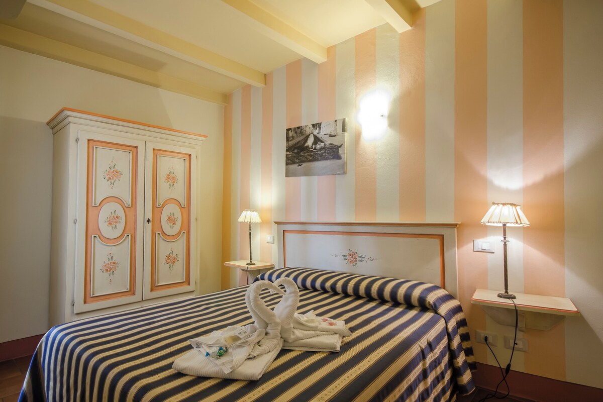 A hotel room with a bed, two bedside lamps, a headboard with a floral design, a wall-mounted picture, and a wardrobe with decorative panels.
