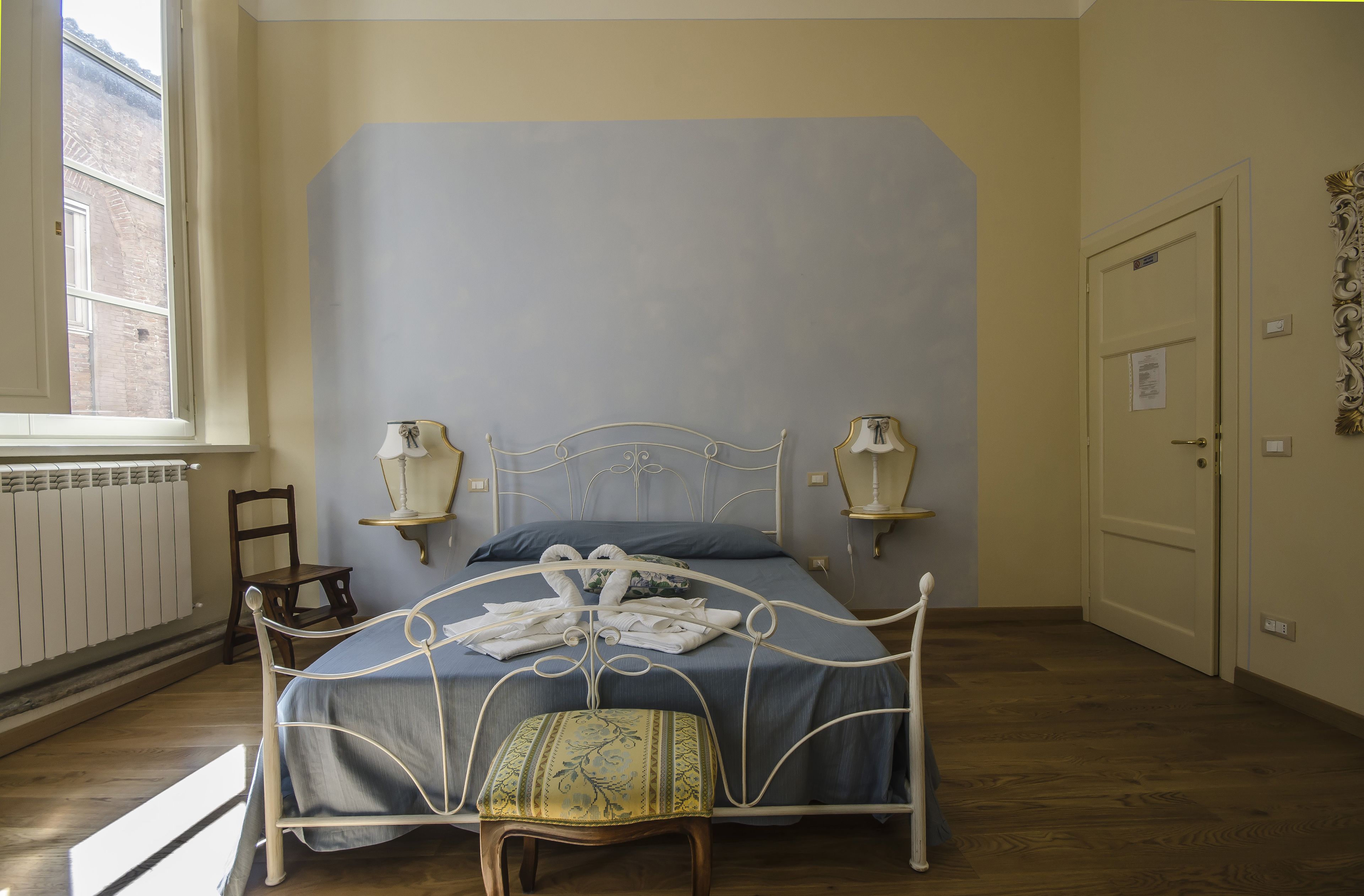 A bedroom with a bed, two bedside lamps, a window, a radiator, a door, and a chair.