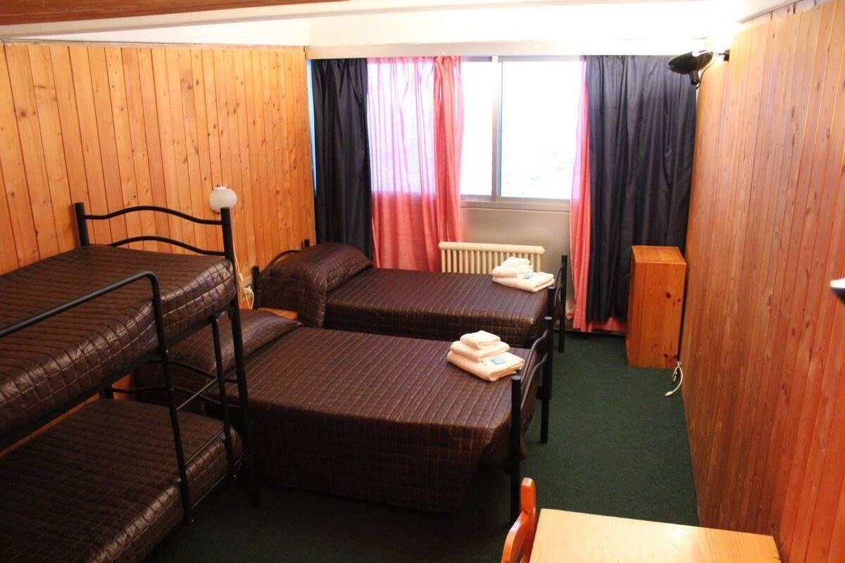 A dormitory room with bunk beds, a radiator, a wooden cabinet, and a window with curtains.