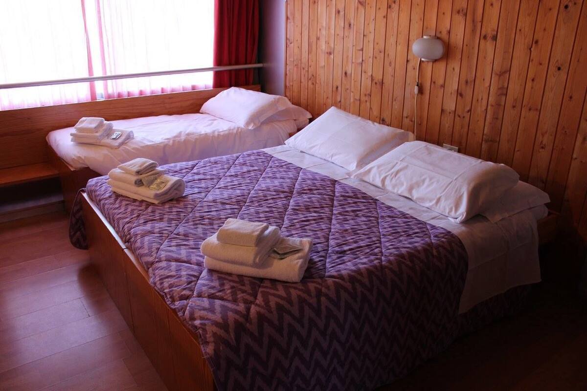 A hotel room with two beds, wooden walls, a window with curtains, and a wall-mounted lamp.