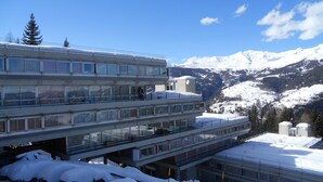 A multi-story building with a snow-covered roof and a view of snow-capped mountains in the background. - Hotel Sole Alto (Mezzana)