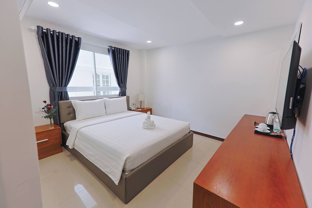Quarto de hotel com uma cama, uma escrivaninha de madeira, uma televisão e uma janela com cortinas.