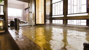 A traditional Japanese onsen with a stone bath, tiled walls, and large windows.