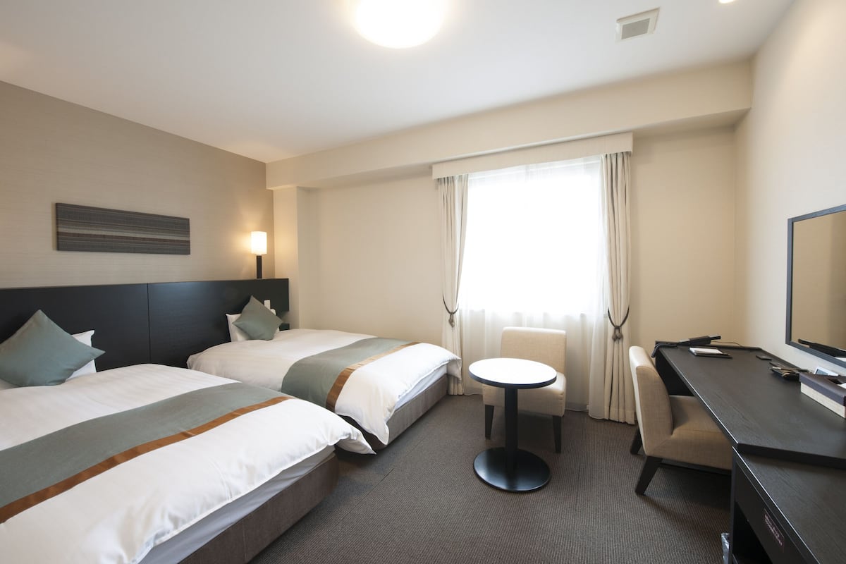 A hotel room with two beds, a round table, a desk, and a large window with curtains.