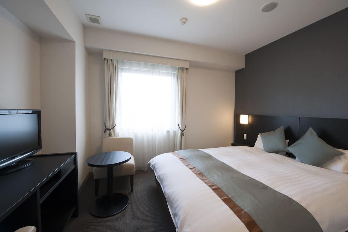 A hotel room with a bed, a TV, a small table, and a window with curtains.