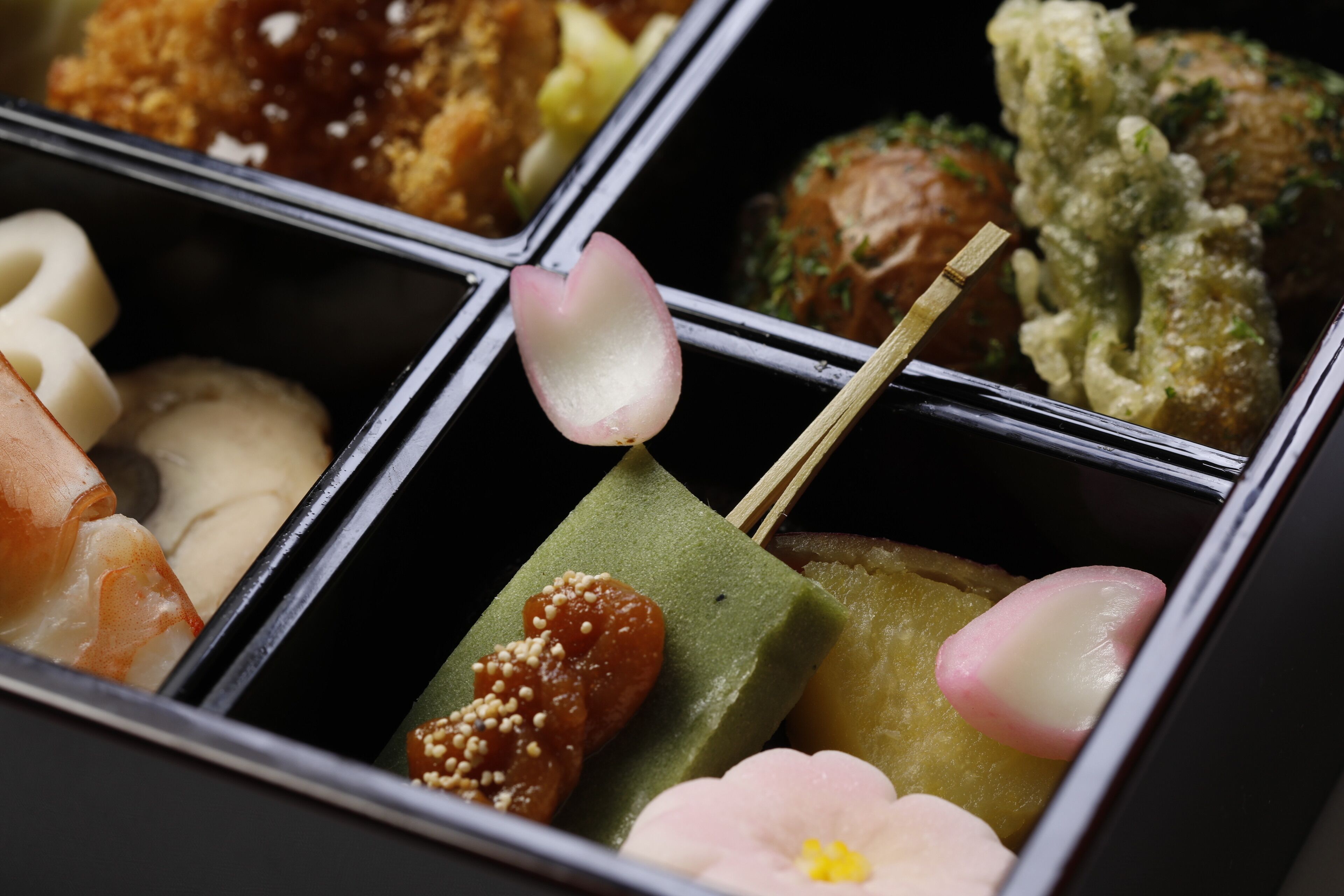 A close-up of a bento box with various food items, including sushi, pickled vegetables, and a piece of green cake with sesame seeds.