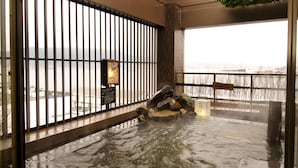 A hot spring area with a stone bath surrounded by a wooden fence, a signboard, and a view of a snowy landscape through the fence.