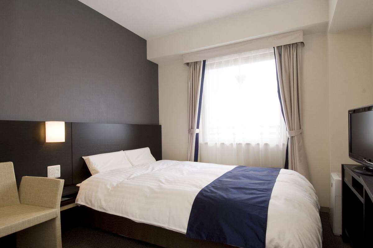A hotel room with a bed, a chair, a TV, and a window with curtains.