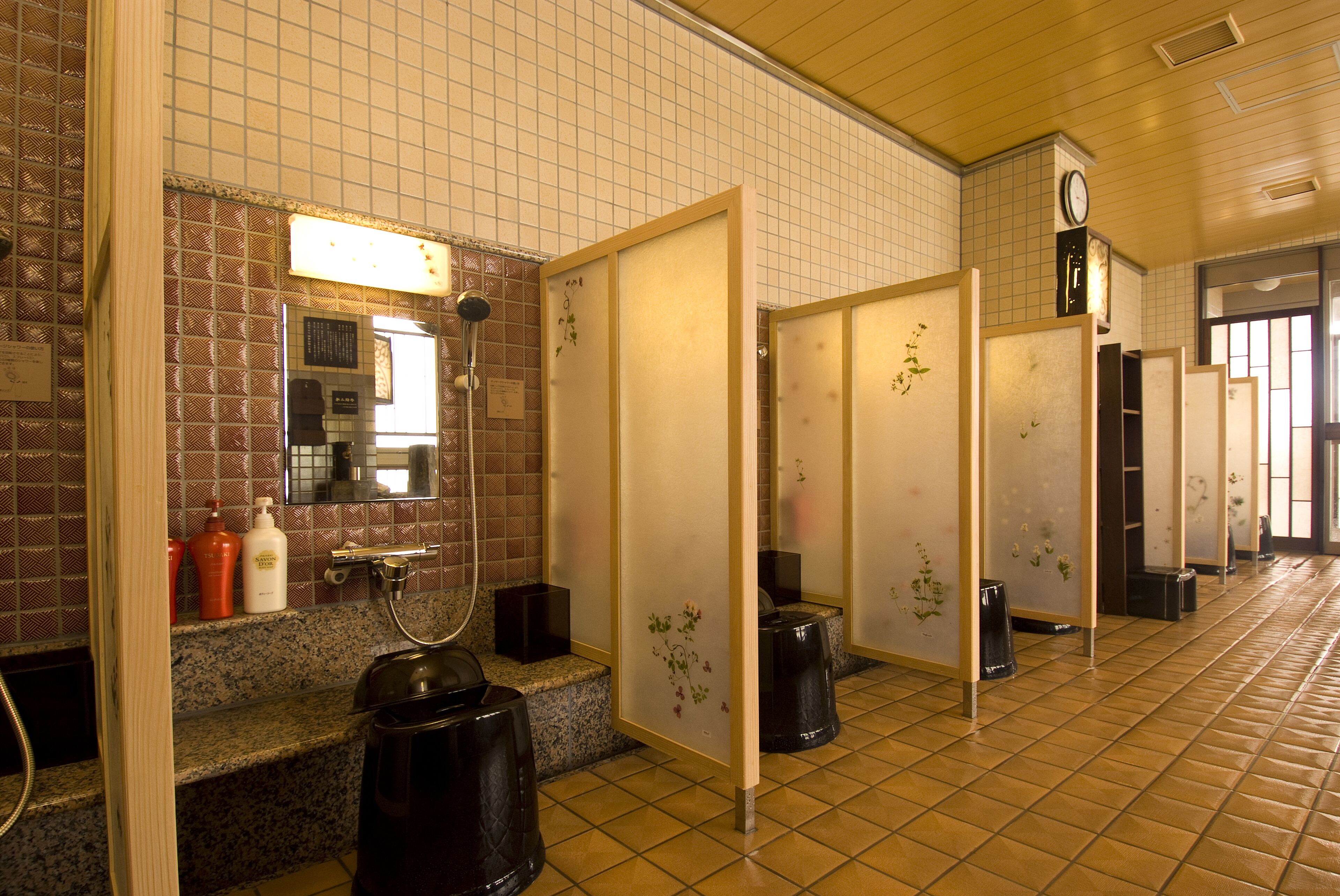 A public restroom with tiled walls, a mirror, a clock, and a trash can.