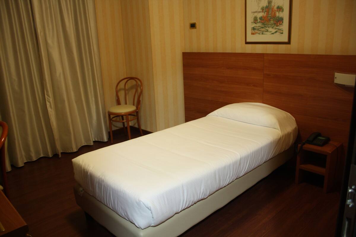 A single bed with a wooden headboard, a chair, a bedside table with a phone, and a curtain.