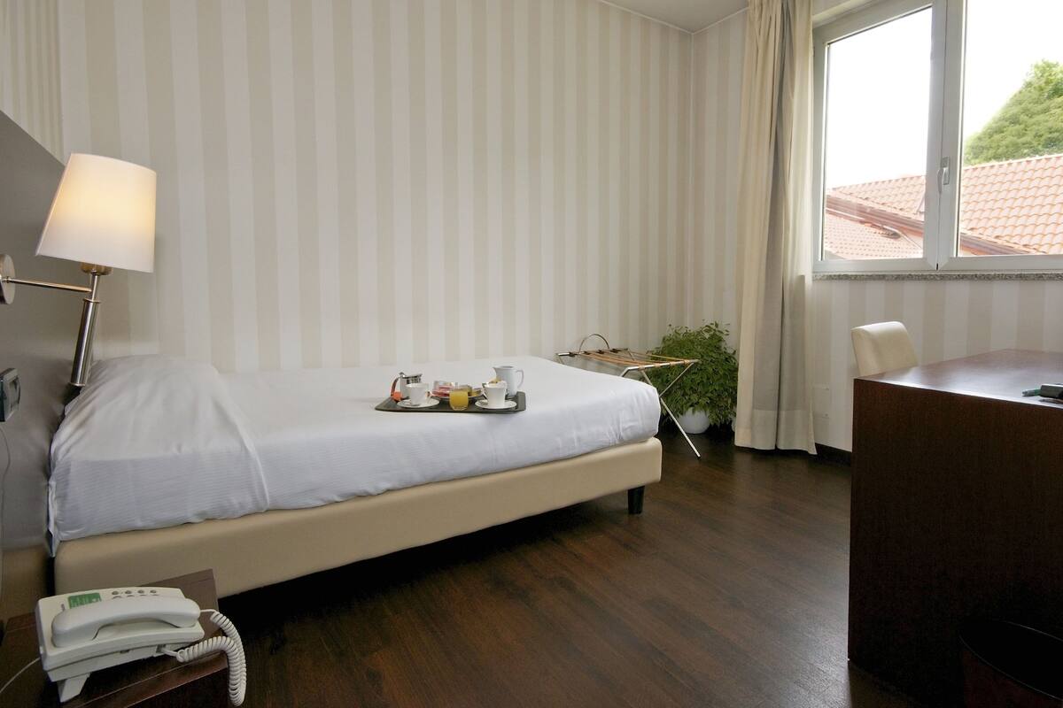 A hotel room with a bed, bedside table, lamp, desk, and a window with curtains.