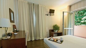 Habitación de hotel con cama, escritorio con lámpara, televisor montado en la pared y ventana con vistas a árboles.