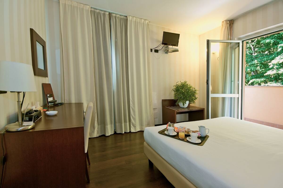 A hotel room with a bed, a desk with a lamp, a TV mounted on the wall, and a window with a view of trees.