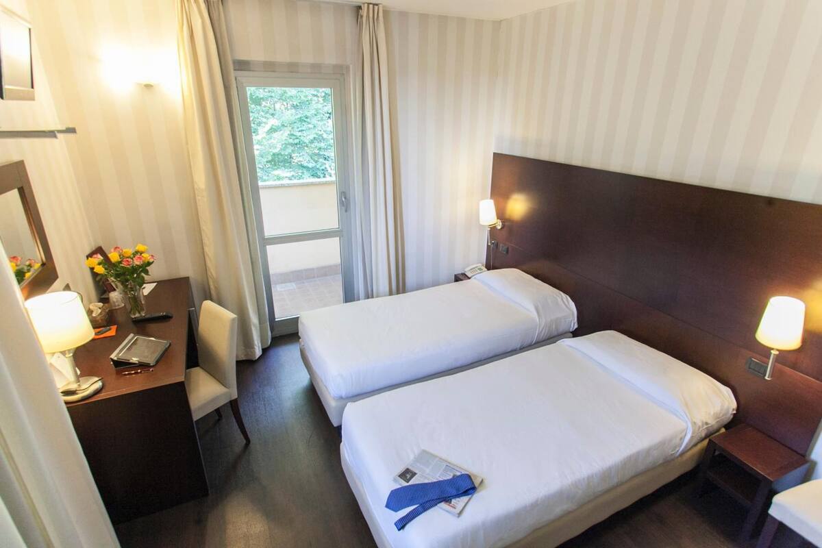 A hotel room with two single beds, a desk with a lamp, a chair, and a window with curtains.