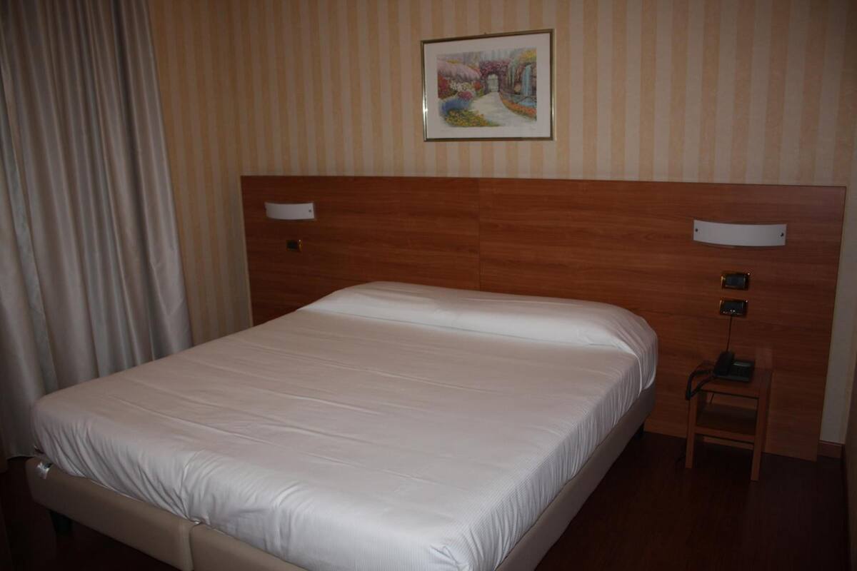 A hotel room with a large bed, wooden headboard, a small bedside table with a lamp, and a painting on the wall.