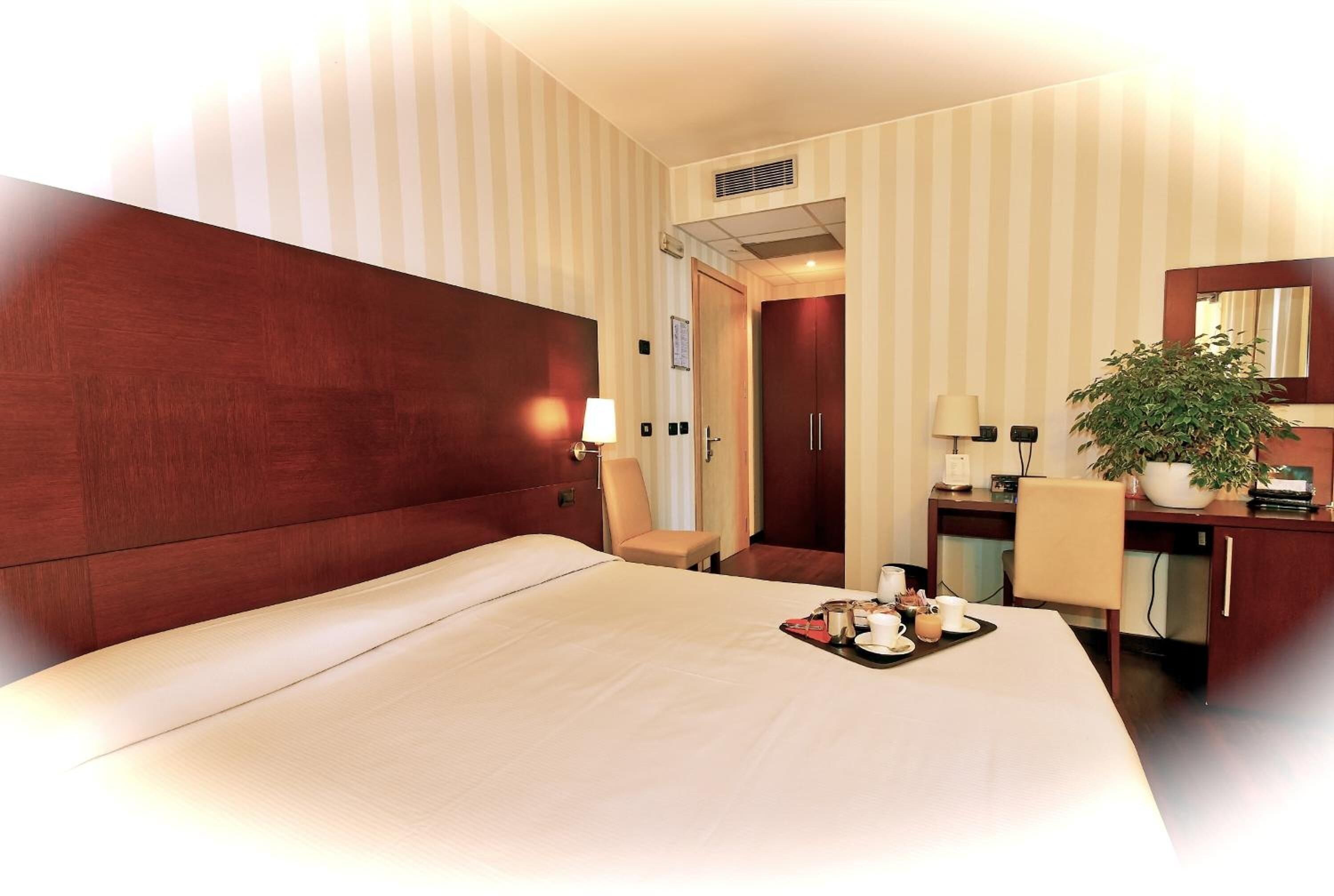 Una habitación de hotel con una cama grande, un escritorio con silla, una planta en maceta y un espejo.