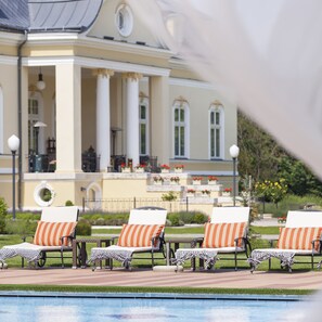 A poolside area with lounge chairs and a large building in the background. - Hotel Château Amade (Vrakun)