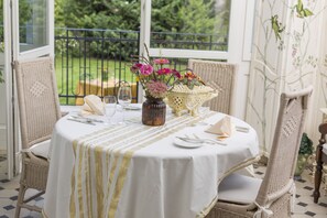 A round dining table with a white tablecloth and a striped table runner. There are wicker chairs around the table, a vase with flowers, and a decorative basket. - Hotel Château Amade (Vrakun)