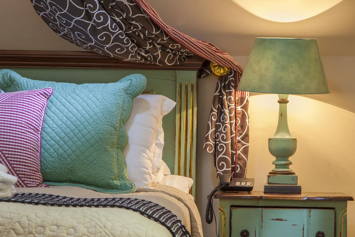 Quarto com cama, almofadas, mesa de cabeceira com abajur e uma cadeira verde.