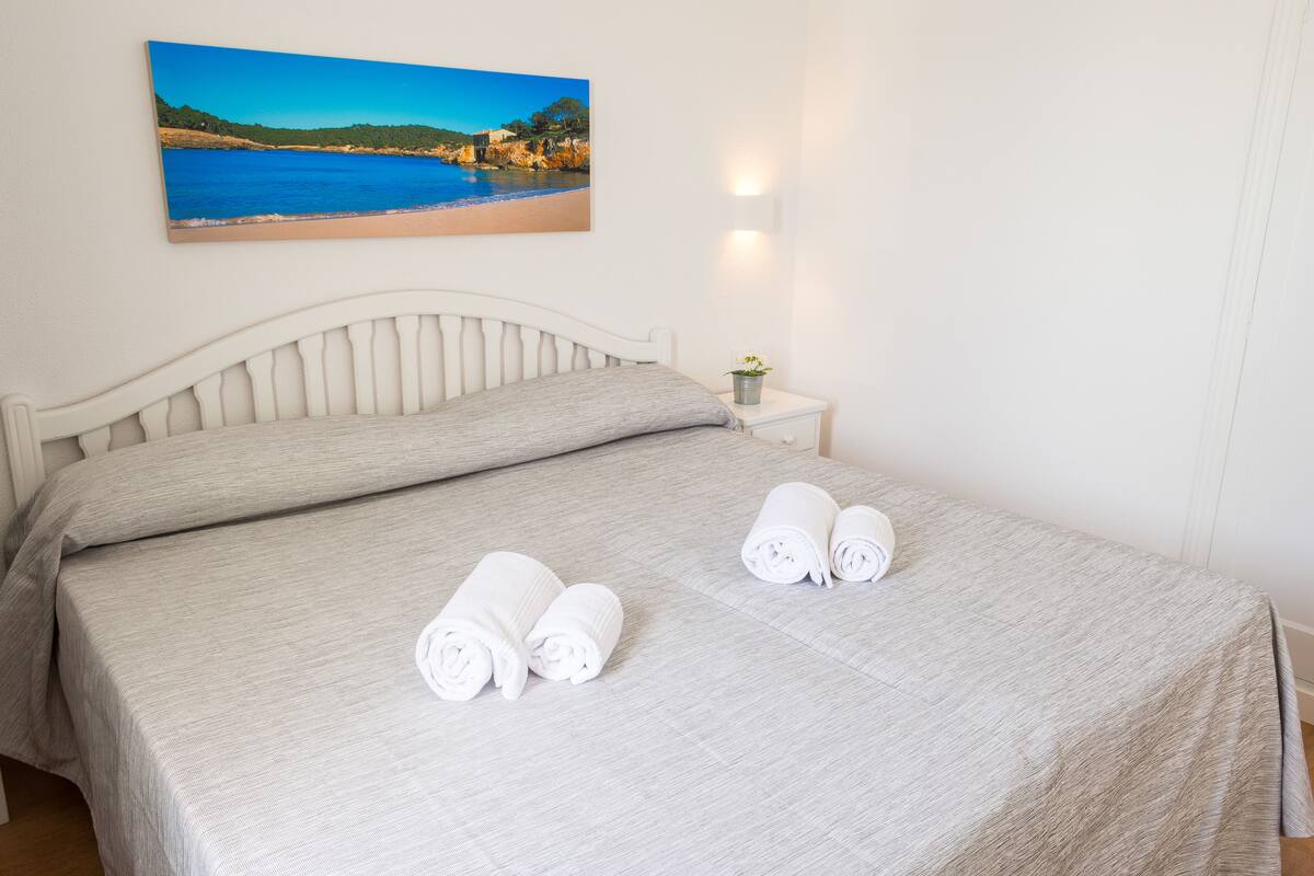 A neatly made bed with a light-colored bedspread and two rolled towels on the bed.