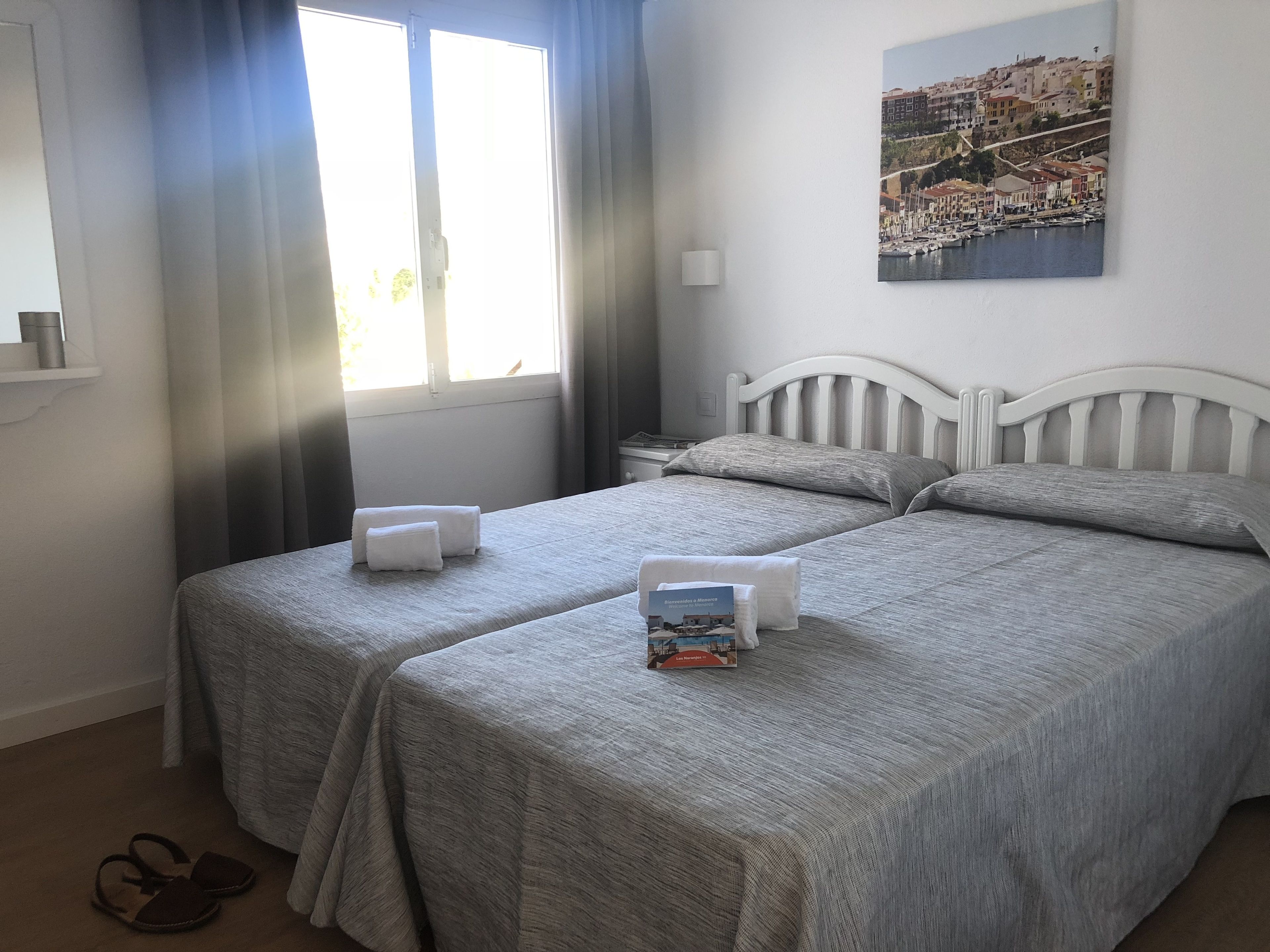 Hotelkamer met twee bedden, een raam met gordijnen, een nachtkastje, een lamp en een schilderij van een kustplaatsje.