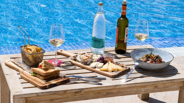 Una mesa junto a la piscina con comida, bebidas y una botella de vino.