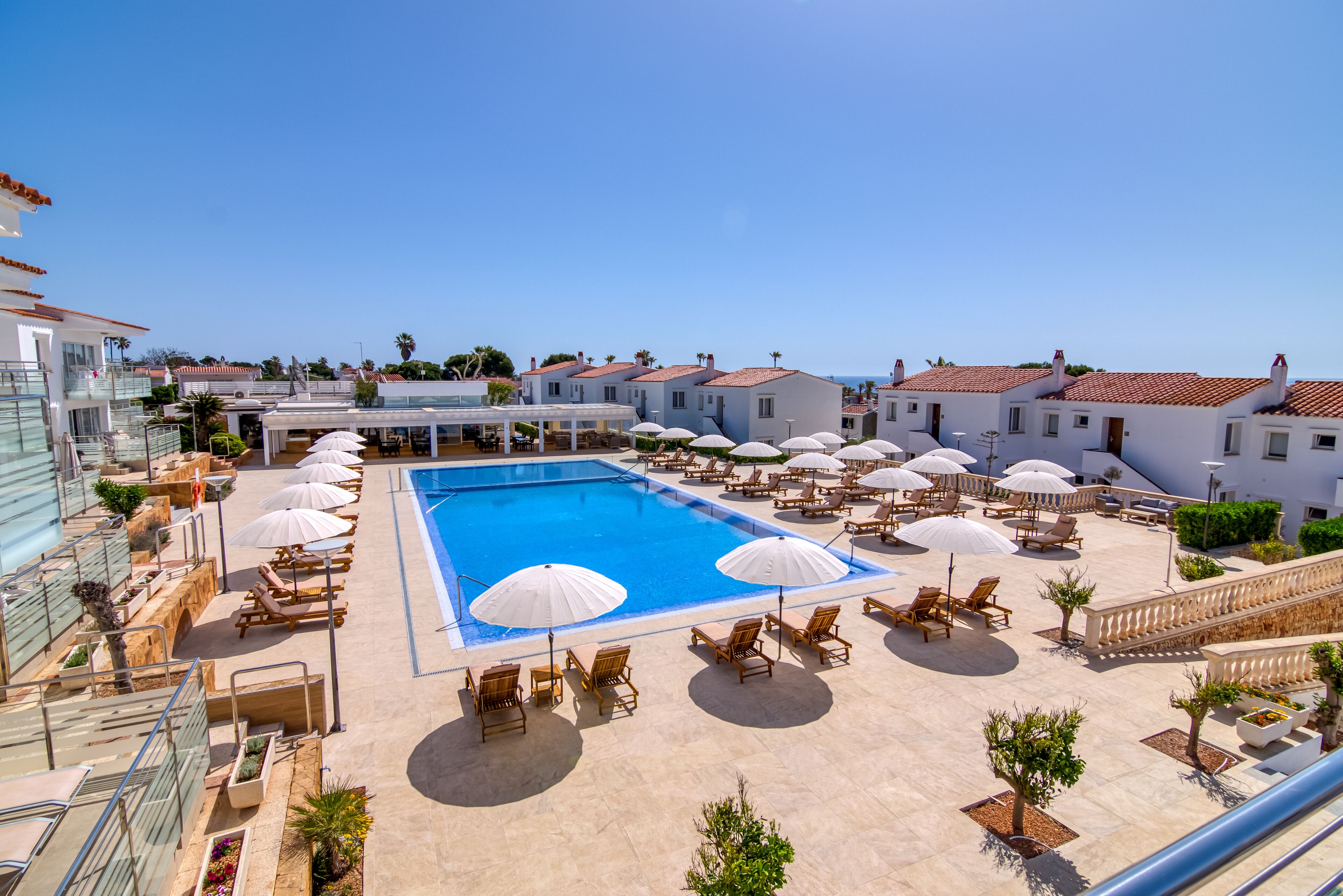 A resort pool area with sun loungers and umbrellas, surrounded by buildings with red-tiled roofs.