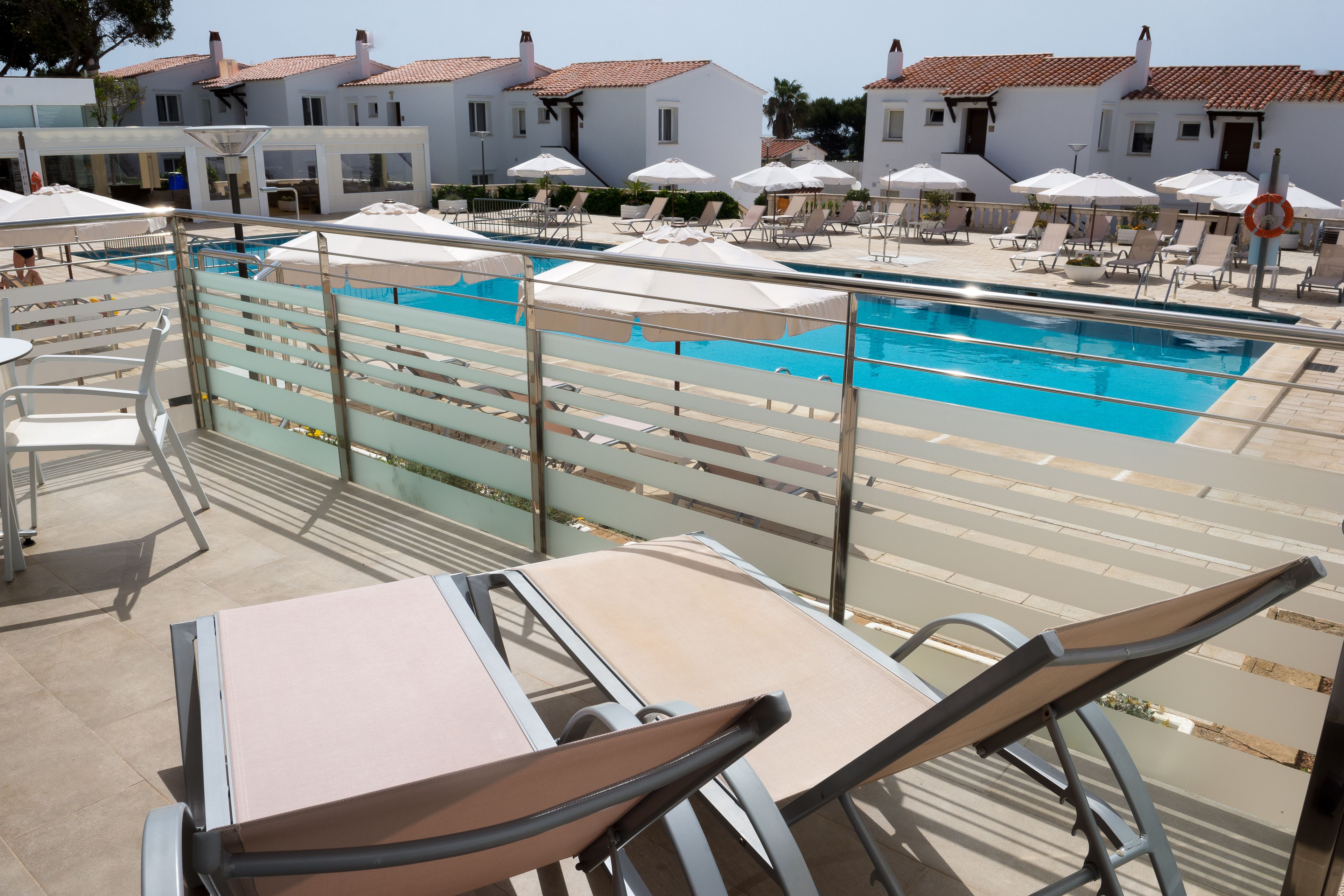 A poolside area with lounge chairs and umbrellas, a pool, and white buildings with red-tiled roofs in the background.