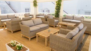 Un amplio espacio al aire libre con mobiliario de mimbre y una mesita de centro, con ventanas grandes y vistas a edificios al fondo.
