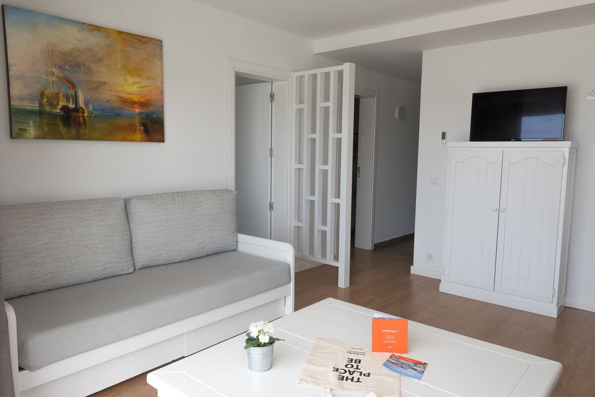 A living room with a grey sofa, a coffee table with a book and a flower vase, a TV on a cabinet, and a painting on the wall.