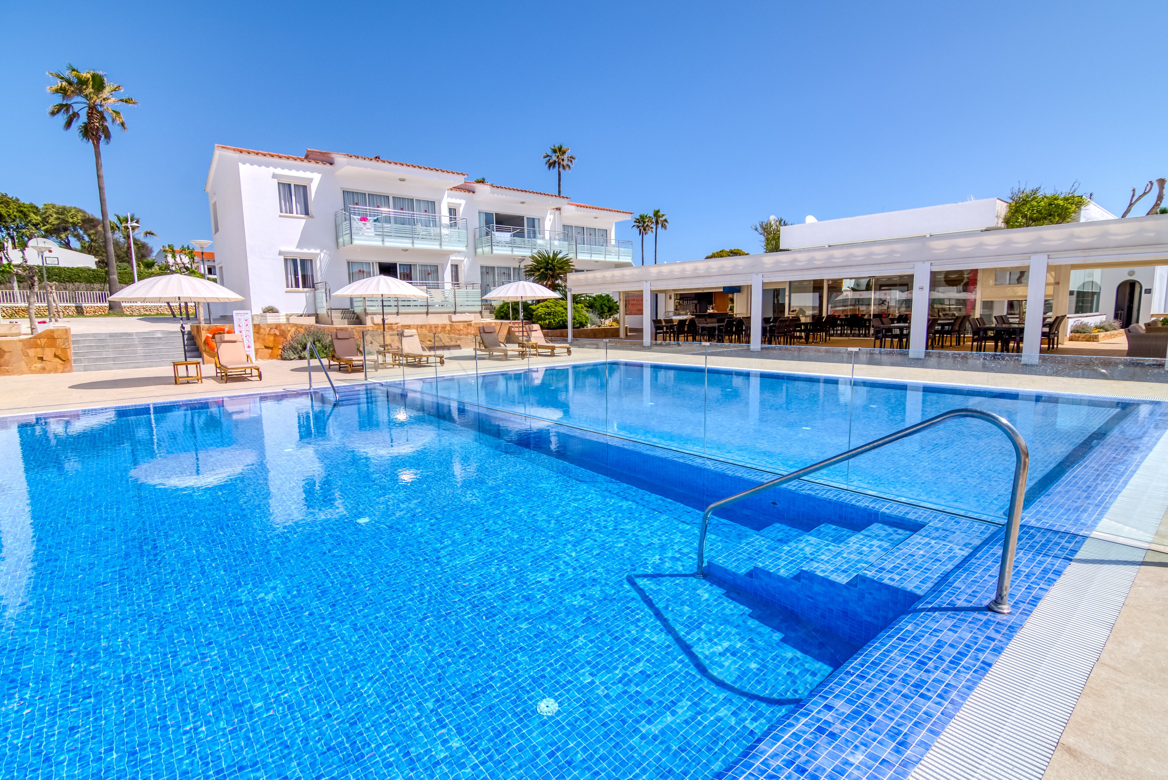 A swimming pool with lounge chairs and umbrellas, a covered outdoor seating area, and a building with balconies.