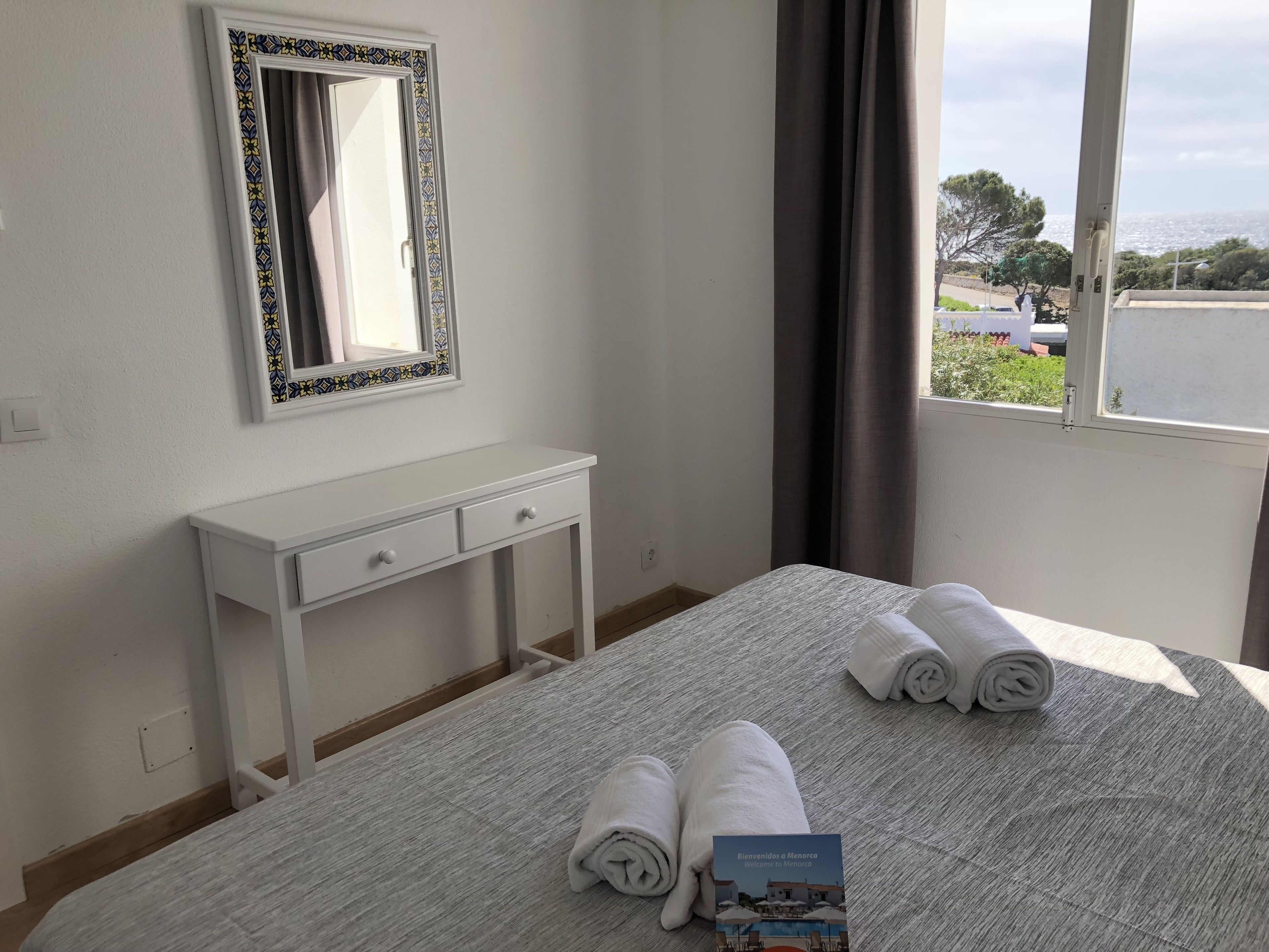 Habitación de hotel con cama, mesita de noche con dos cajones, espejo con marco decorativo, ventana con cortinas y una tarjeta de bienvenida sobre la cama.