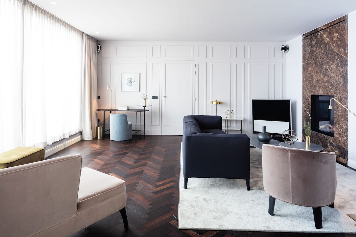 Una sala moderna con piso de madera en espiga oscura, un sofá color beige, un sillón oscuro y un reposapiés redondo. Un televisor de pantalla plana está montado en la pared, y se observa una chimenea al fondo.