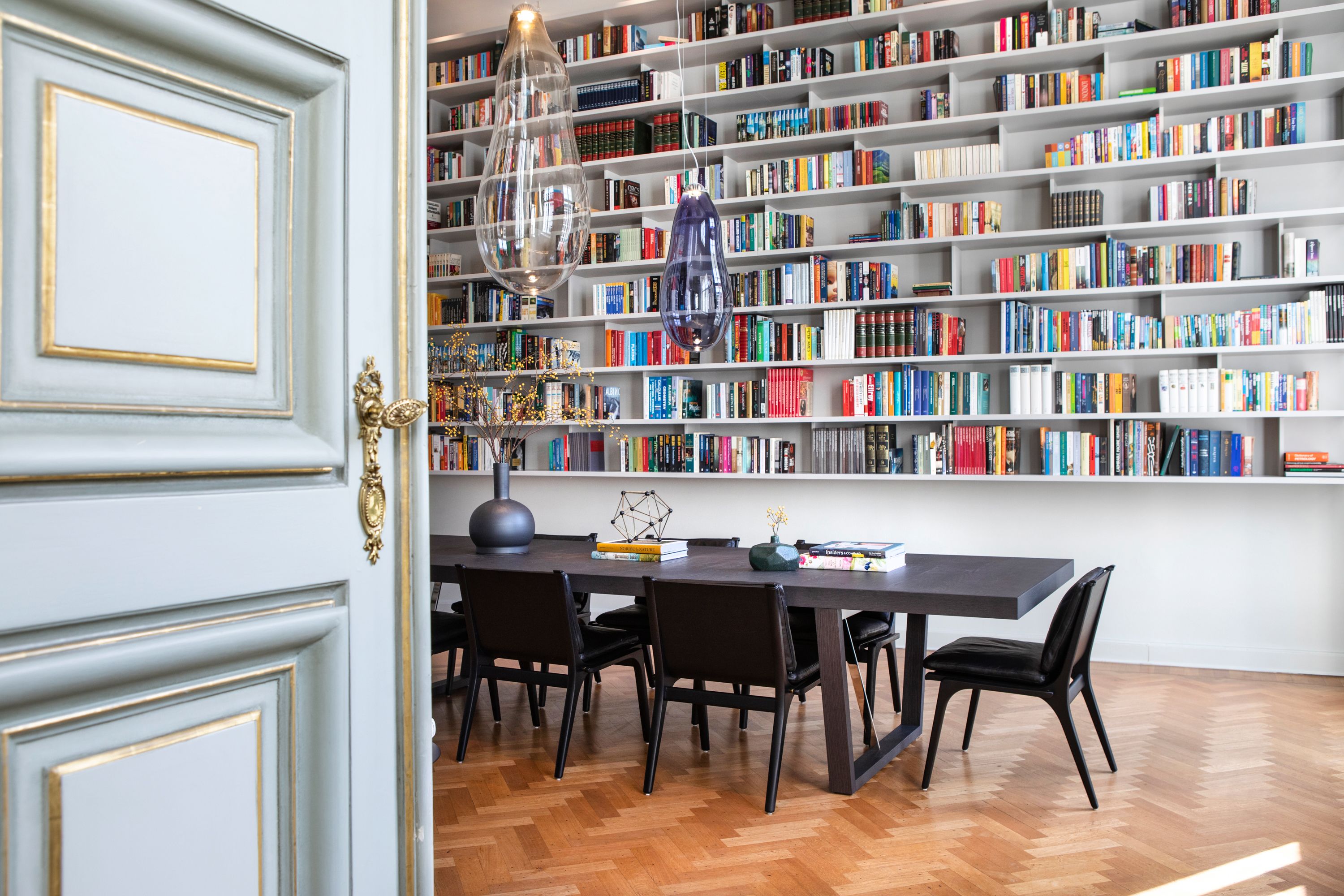 A room with a wooden herringbone floor, a dark wooden table with four chairs, and a bookshelf filled with books.