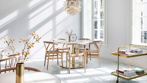 A bright room with a round wooden table, chairs, a gold-framed hanging light, and a window with blinds.