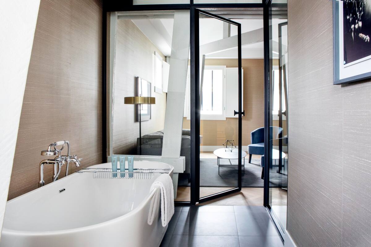 Un baño moderno con una bañera independiente, un box de ducha de vidrio y una zona de estar visible con una mesa redonda y sillas.