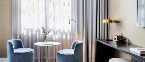 A modern hotel room with two blue armchairs, a small round table, a floor lamp, a desk with a radio, and a large window with sheer curtains.