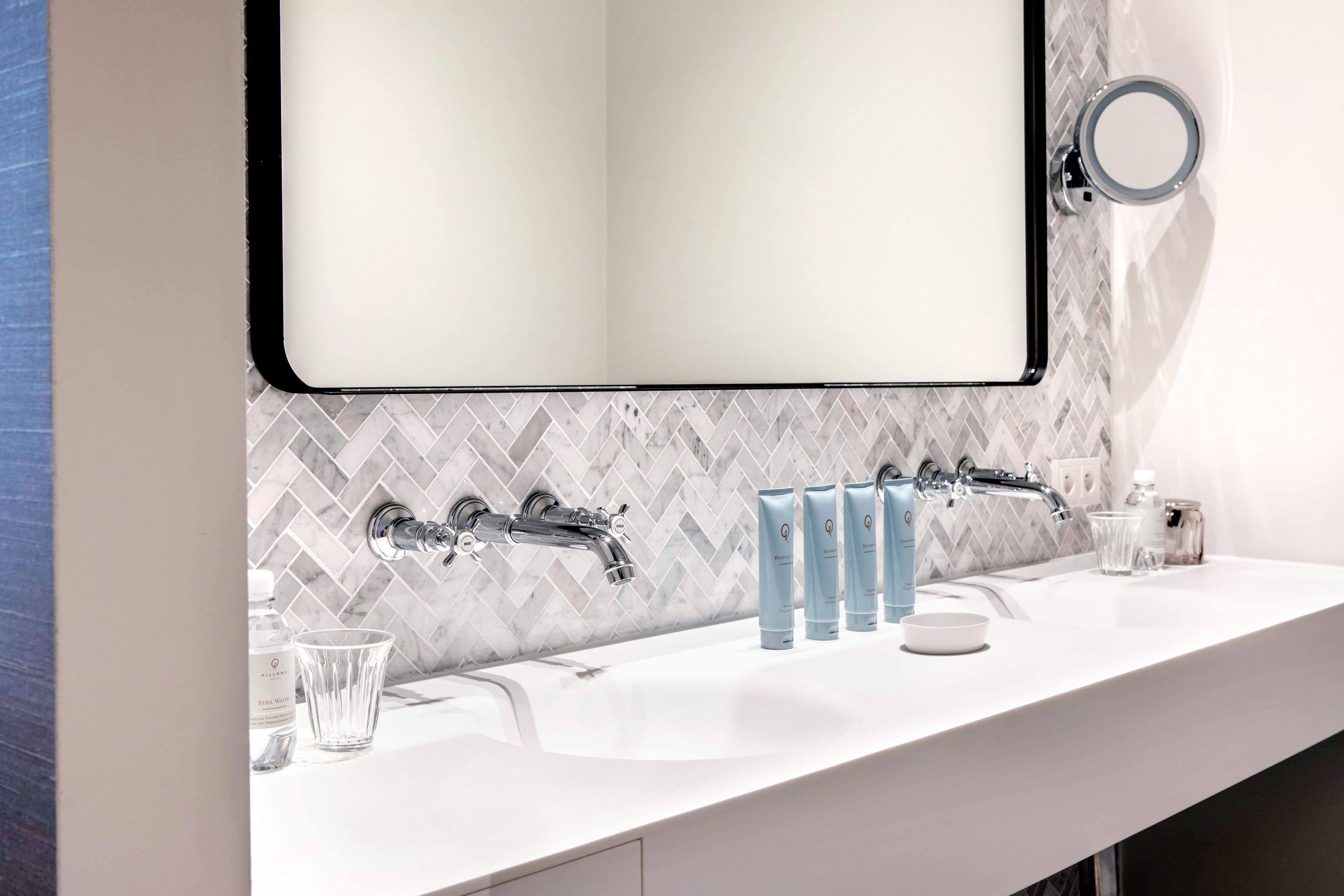 A modern bathroom with a white sink, two faucets, and a mirror. There are toiletries on the counter, including a soap dispenser and a glass.