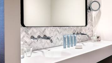 Une salle de bain moderne avec un lavabo blanc, deux robinets et un miroir. On y trouve des articles de toilette sur le comptoir, notamment un distributeur de savon et un verre.