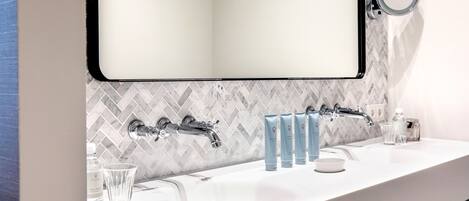 A modern bathroom with a white sink, two faucets, and a mirror. There are toiletries on the counter, including a soap dispenser and a glass.