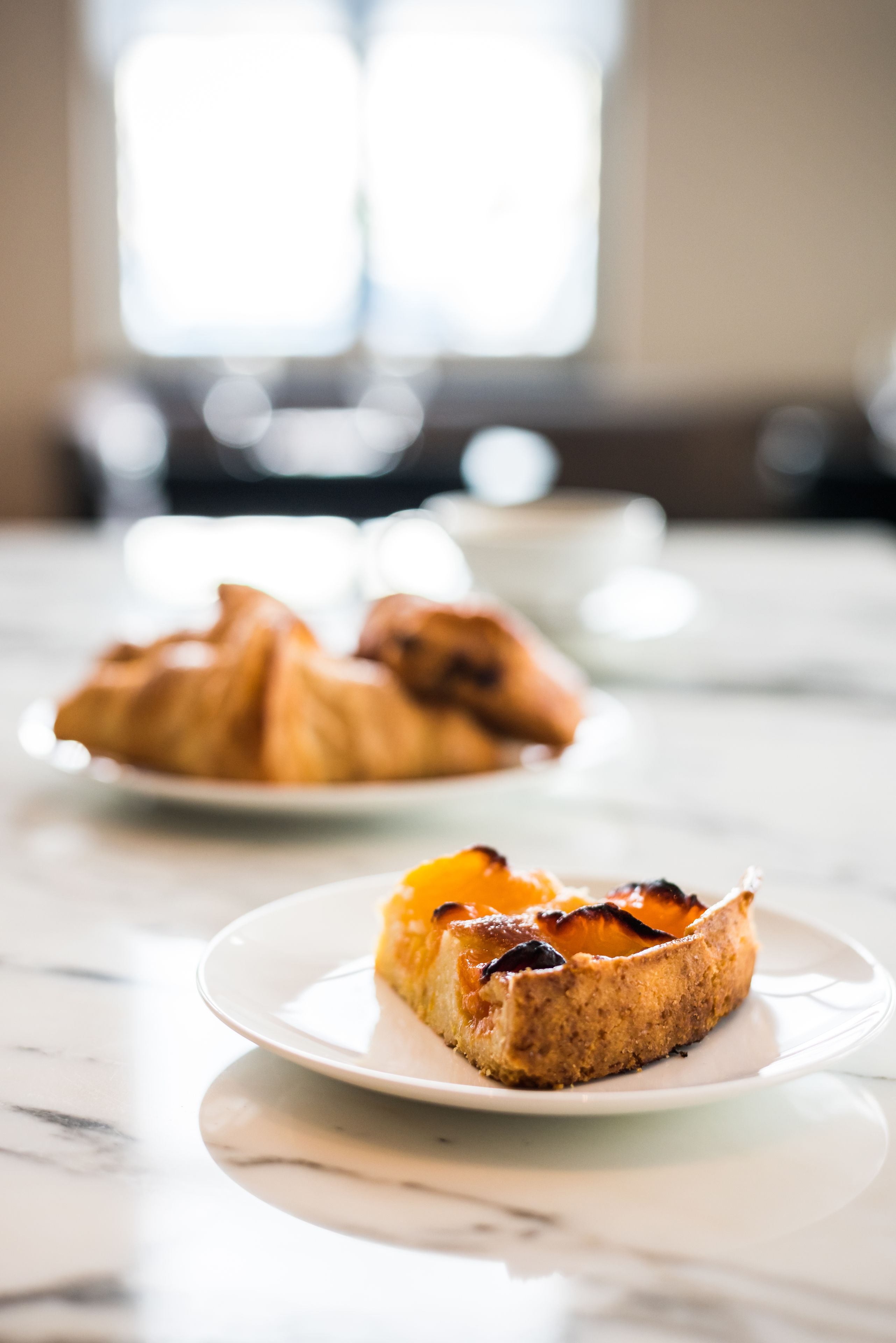 Et bord med to tallerkener mad, hvor den ene har ristet brød og den anden croissanter, i et lyst rum med et vindue i baggrunden.